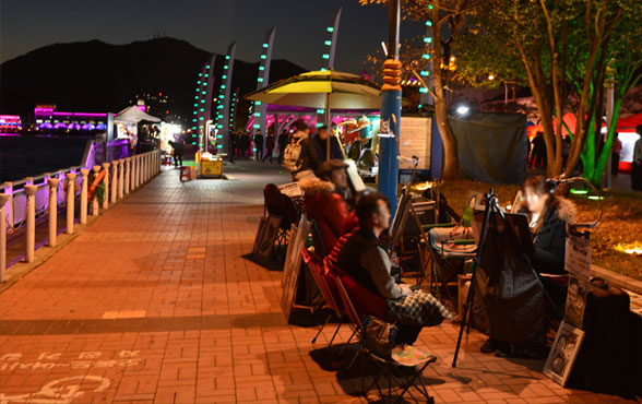 해양공원 밤 풍경으로 좌측에는 자전거길이 보이고 우측에는 앉아있는 노점상인의 모습이 보인다.