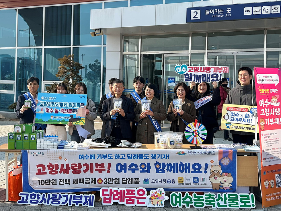 여수에서 고향사랑기부제를 홍보하는 행사로, 여러 사람들이 단체로 모여 기부를 독려하고 있다. 테이블에는 기부 관련 물품과 안내판이 놓여 있다.