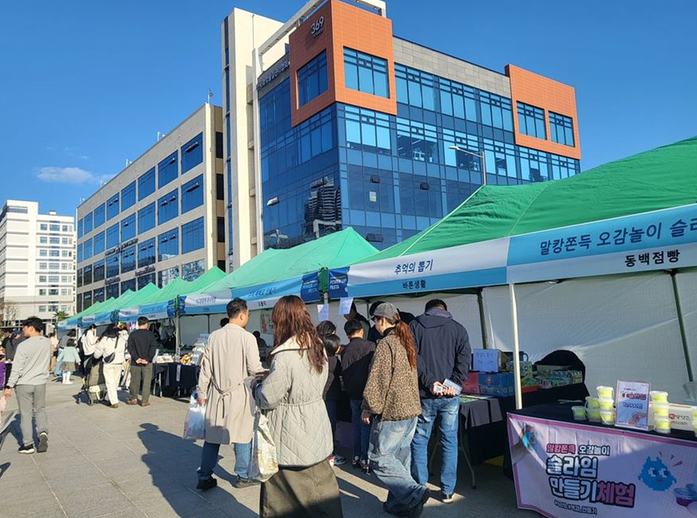 사람들이 야외에서 열린 행사에 참여하고 있으며, 여러 개의 초록색 천막 아래에서 다양한 물품을 판매하고 있다.