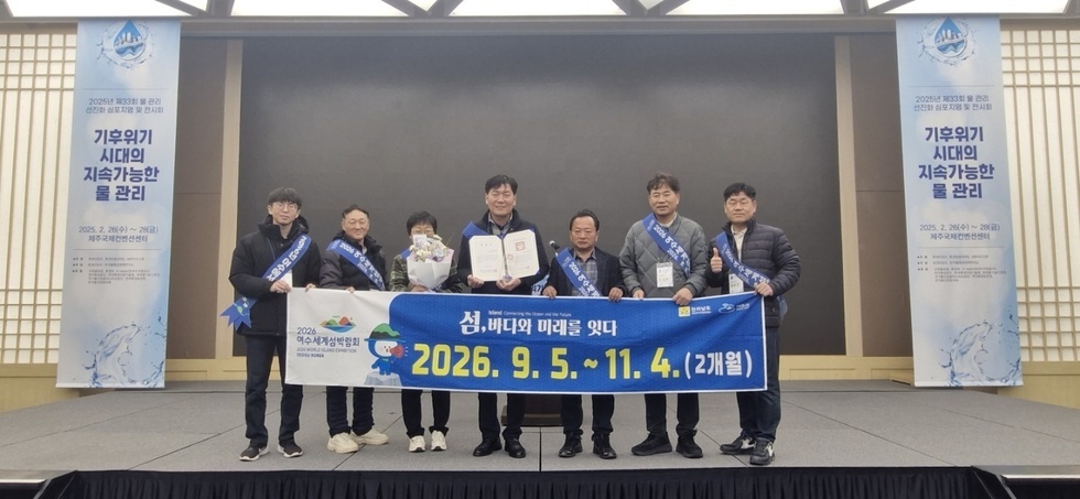 여수세계섬박람회 관련 행사에서 여러 사람들이 기념사진을 찍고 있으며, 가운데 남성이 상장을 들고 있다. 배경에는 행사 일정과 주제가 적힌 현수막이 있다.