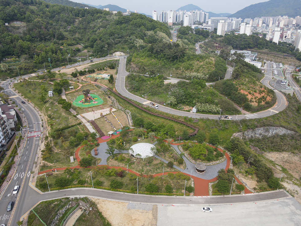 공원 전경이 보이는 사진으로, 다양한 놀이시설과 산책로가 있는 녹지 공간이 펼쳐져 있다. 배경에는 고층 건물이 보인다.