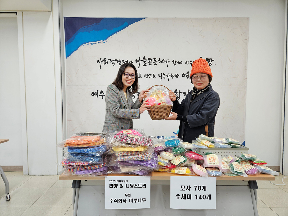 두 사람이 실내에서 기념촬영을 하고 있다. 테이블 위에는 다양한 색상의 손뜨개 모자와 수세미가 정리되어 놓여 있으며, 안내판에는 ‘모자 70개, 수세미 140개’가 적혀 있다. 뒤편 현수막에는 ‘사회적경제와 마을공동체가 함께 키우는 여수의 품격, 공동체로 만드는 지속가능한 여수’라는 문구가 보인다.